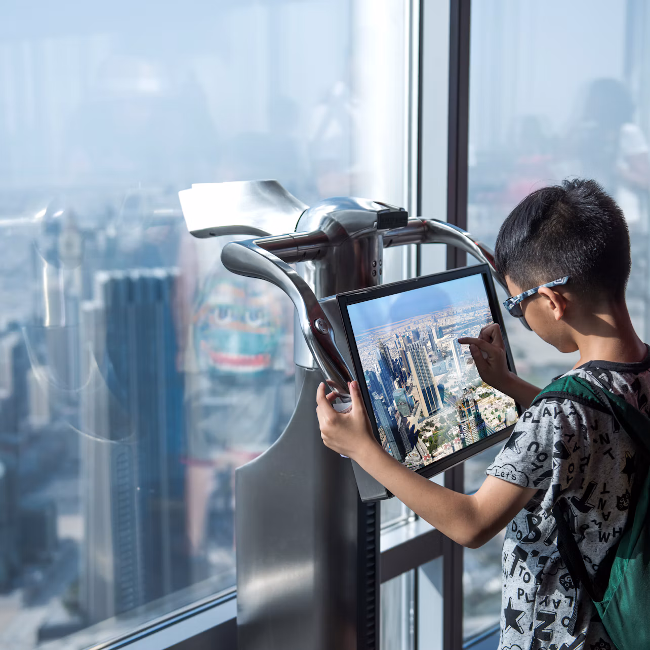 A boy uses a touchscreen display beside a large window with a high-rise cityscape view in the background.