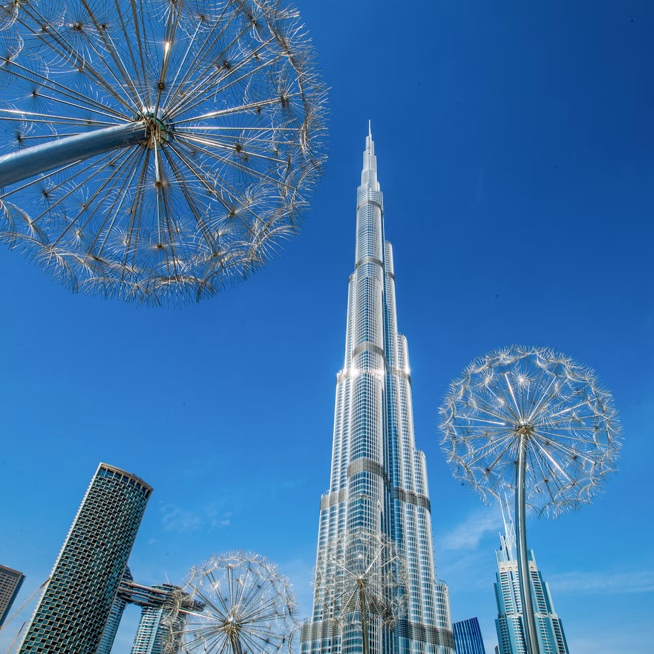 Tall skyscraper against blue sky, surrounded by large metal dandelion sculptures and other modern buildings.