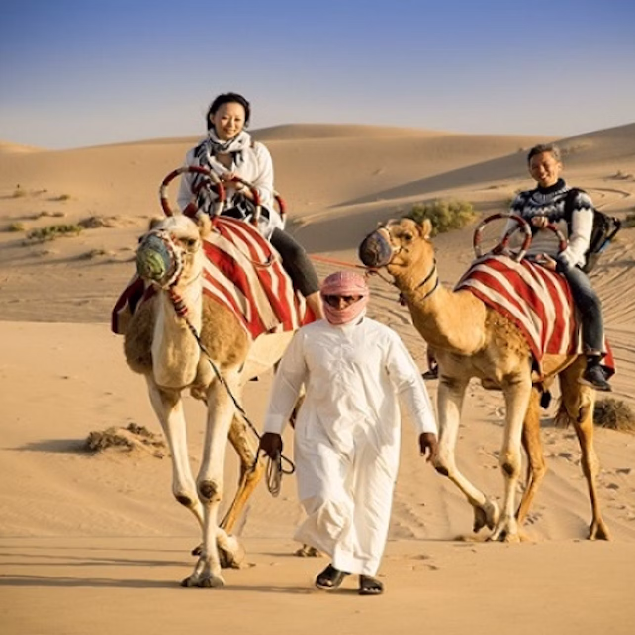Two people riding camels with red and white striped blankets, led by a man in white robes, in a sandy desert setting.