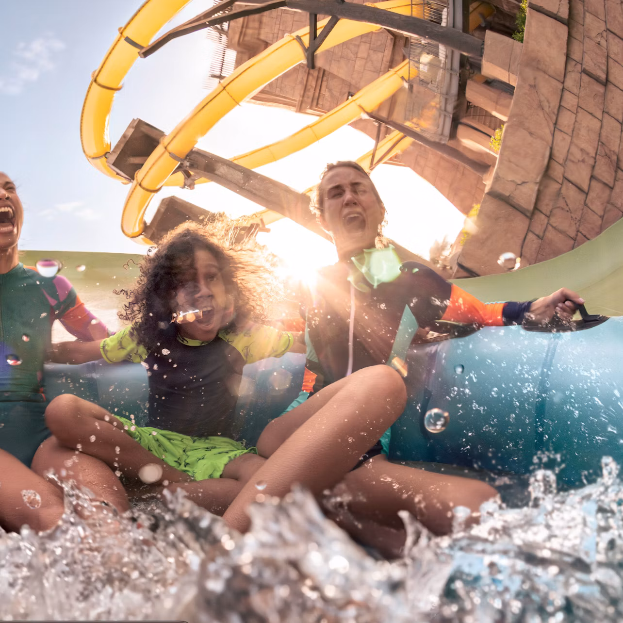 Three people laughing and enjoying a water ride with splashes, against a backdrop of a slide and sunlight.