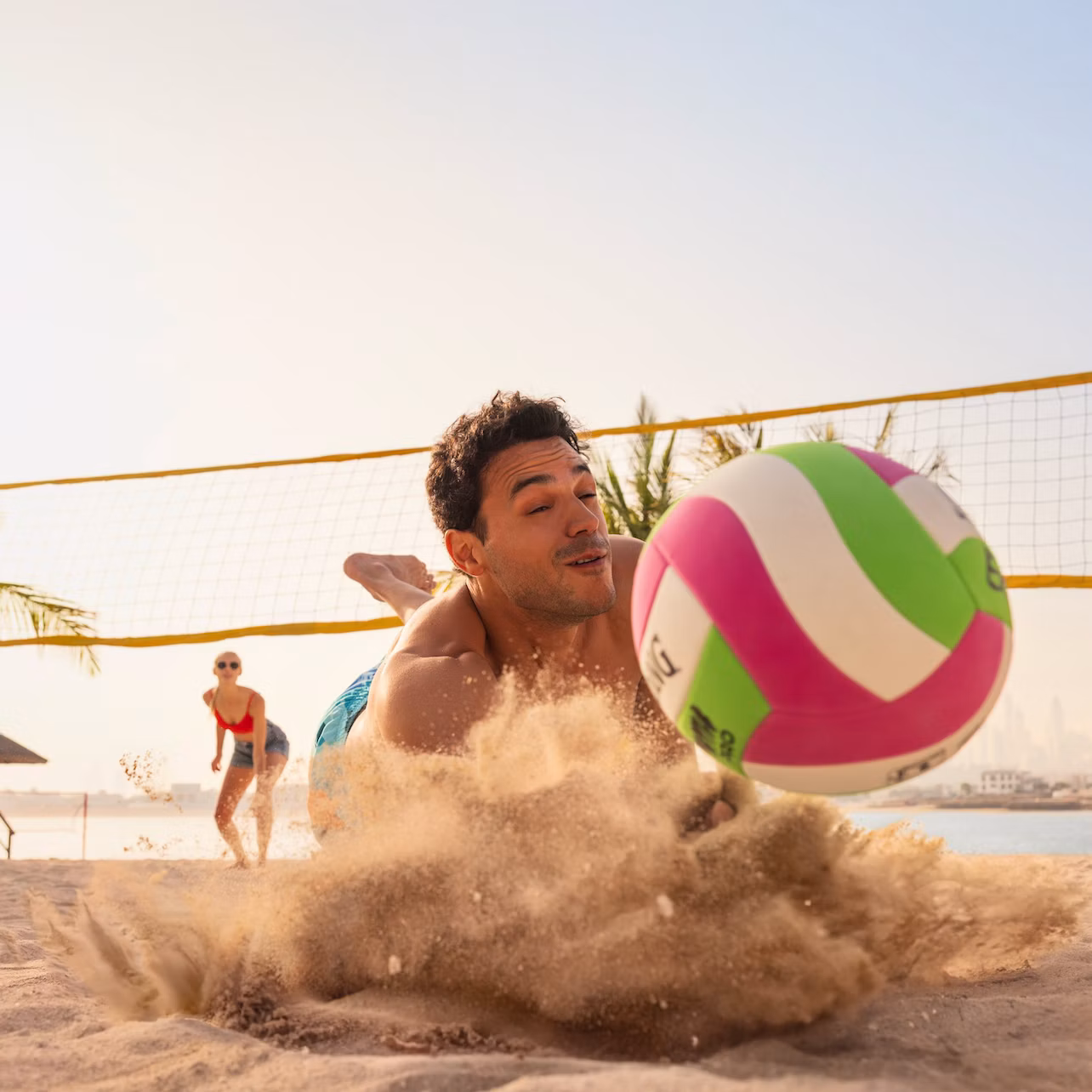 A person dives for a volleyball on a sandy beach, with another person and a volleyball net visible in the background.