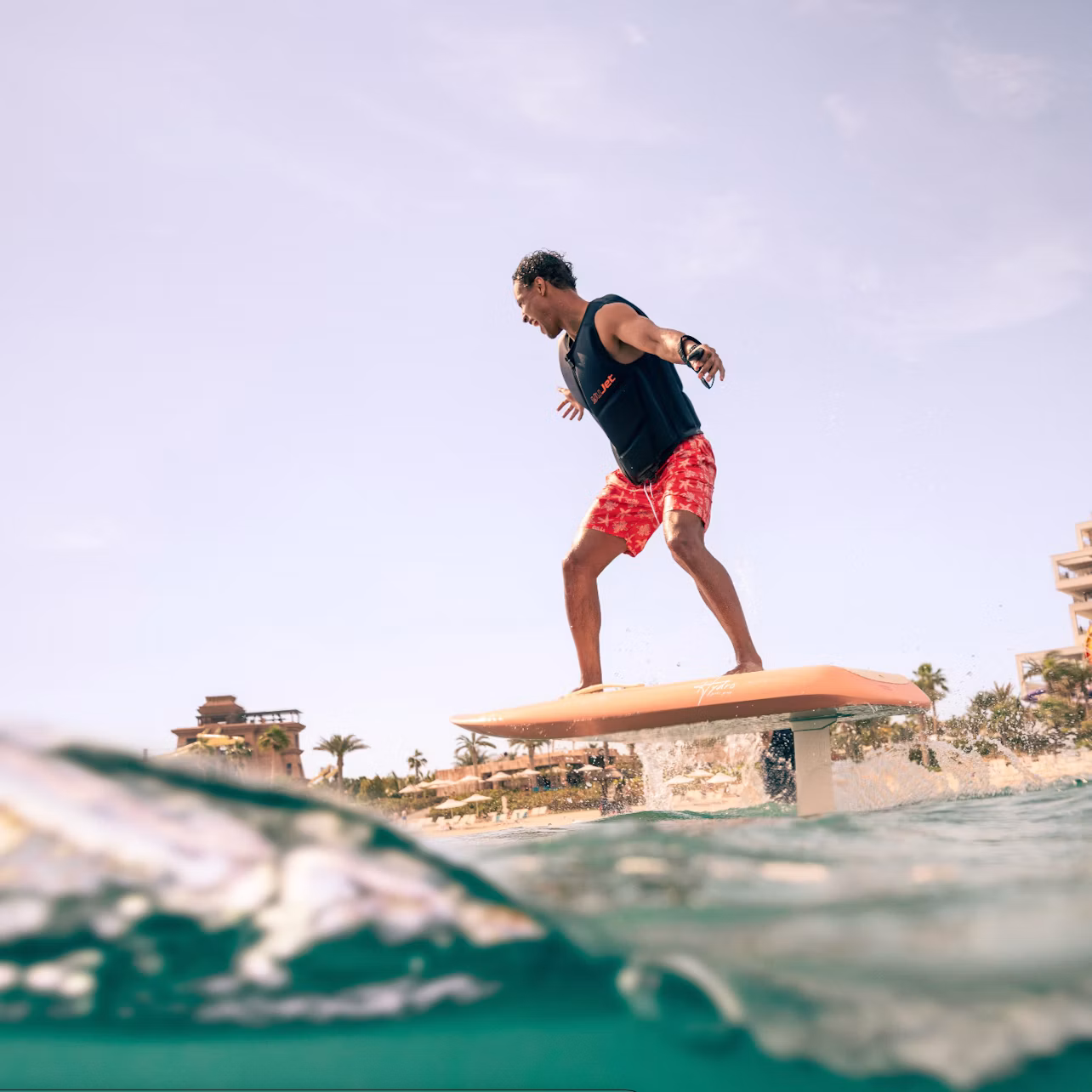 A person balances on a hydrofoil surfboard above water near a beach, with buildings and palm trees visible in the background.