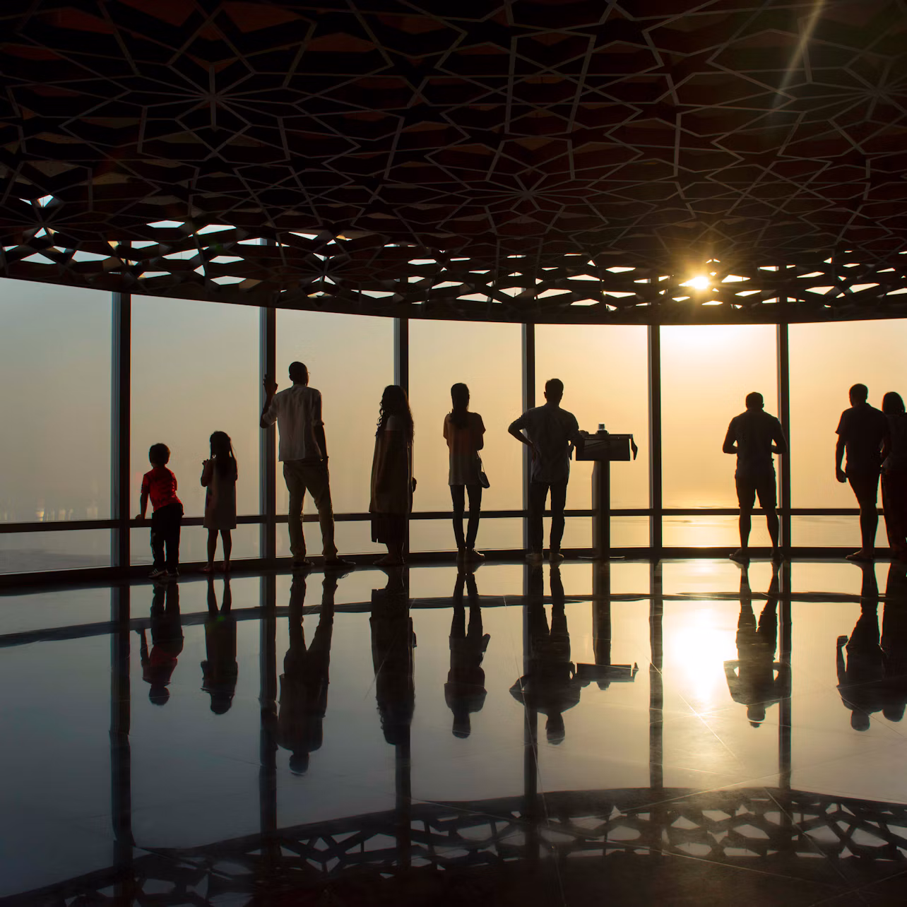 Silhouetted people stand by large windows at sunset, in a room with a patterned ceiling and reflective floor.