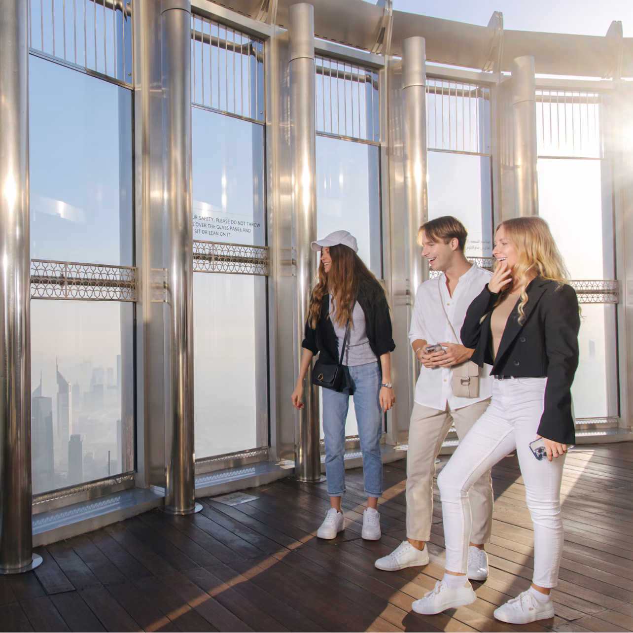 Three people standing on an observation deck with large windows, looking at a city view. They are casually dressed and smiling.