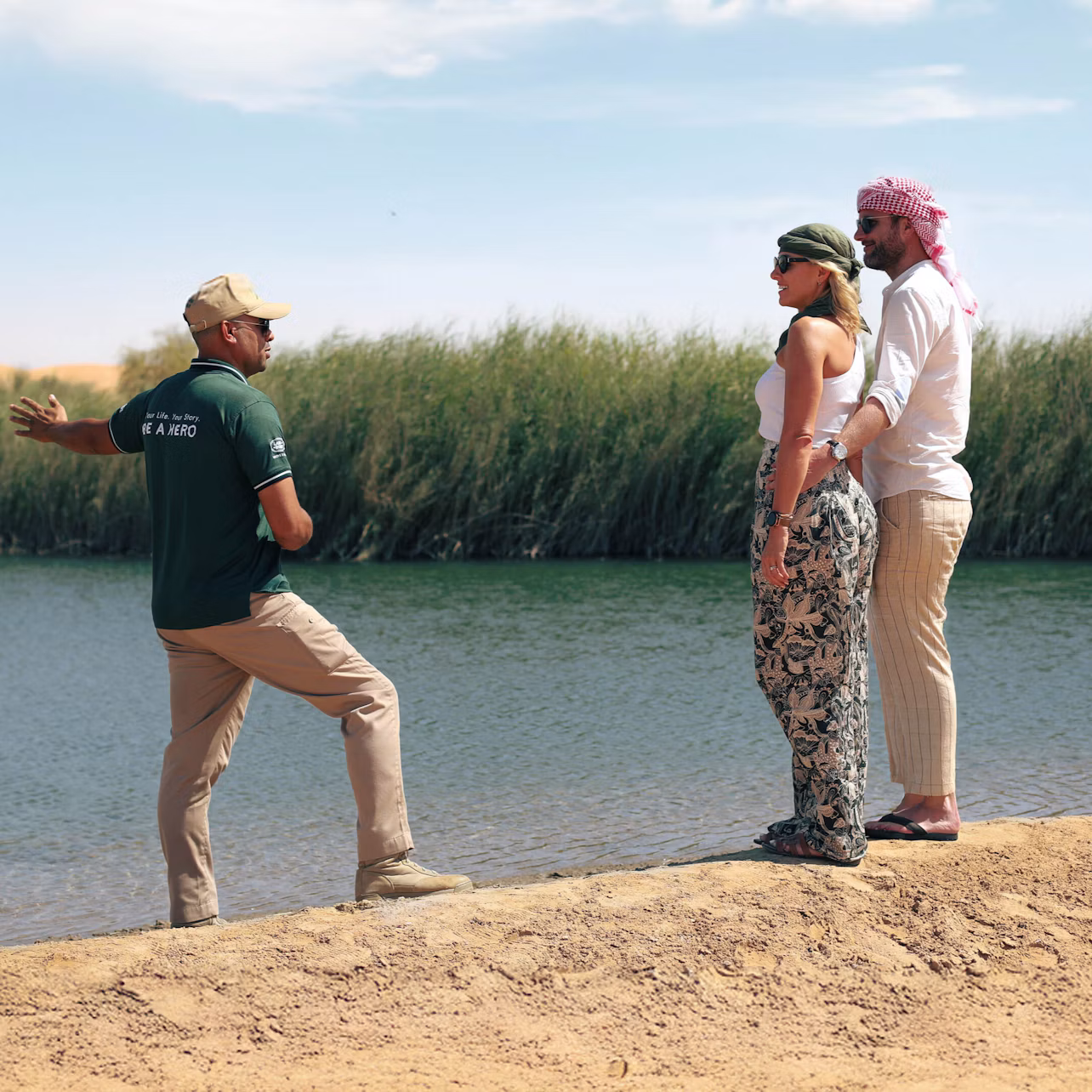 A guide speaks to two tourists near a lush riverbank under a partly cloudy sky.