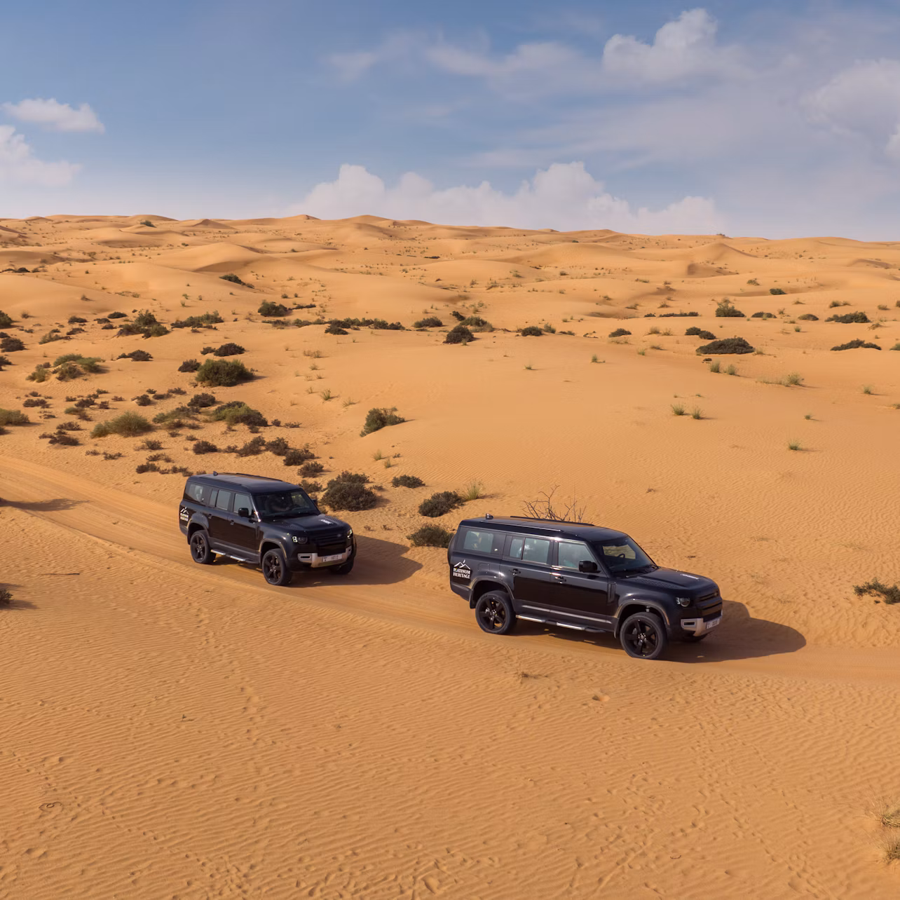 Two black SUVs drive through a vast, sandy desert with small bushes scattered across the landscape under a partly cloudy sky.