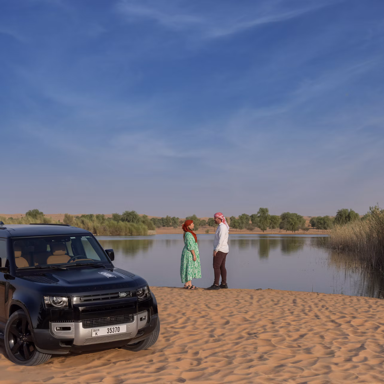 A black SUV parked on sand beside two people standing near a calm waterbody under a clear blue sky.