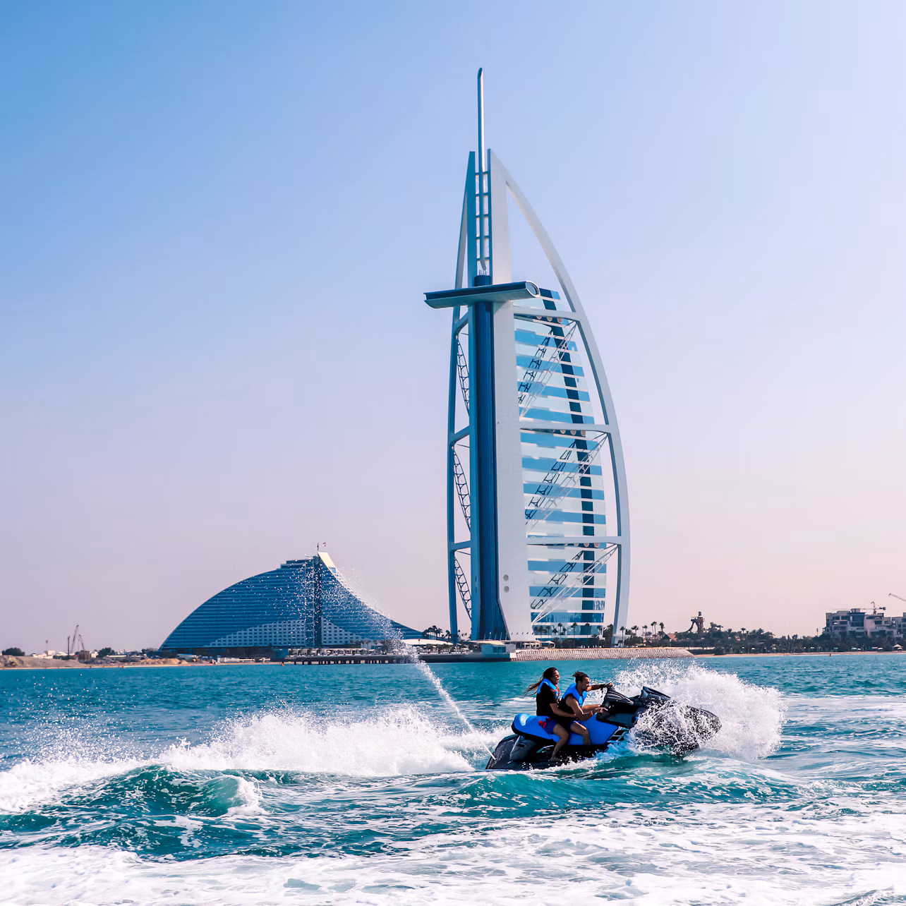 Two people ride a jet ski on blue waters with the Burj Al Arab hotel and another curved building in the background on a clear day.