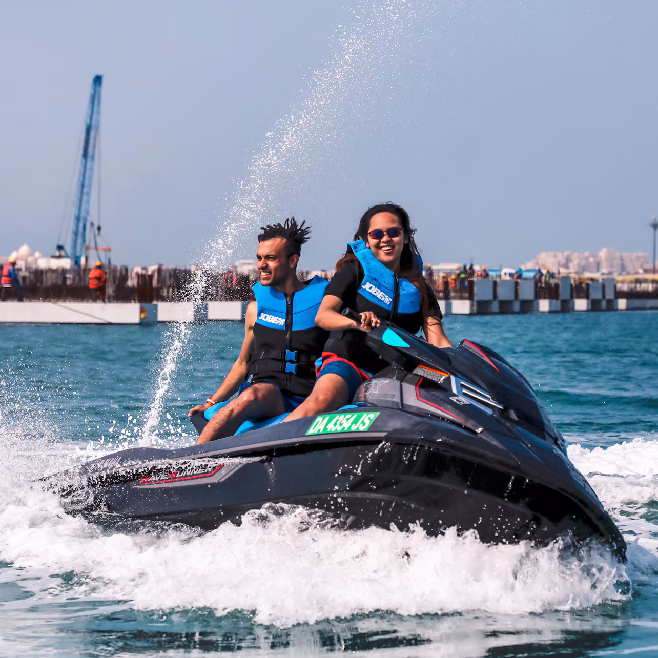 Two people wearing life jackets riding a jet ski on the water with a pier and a crane in the background.