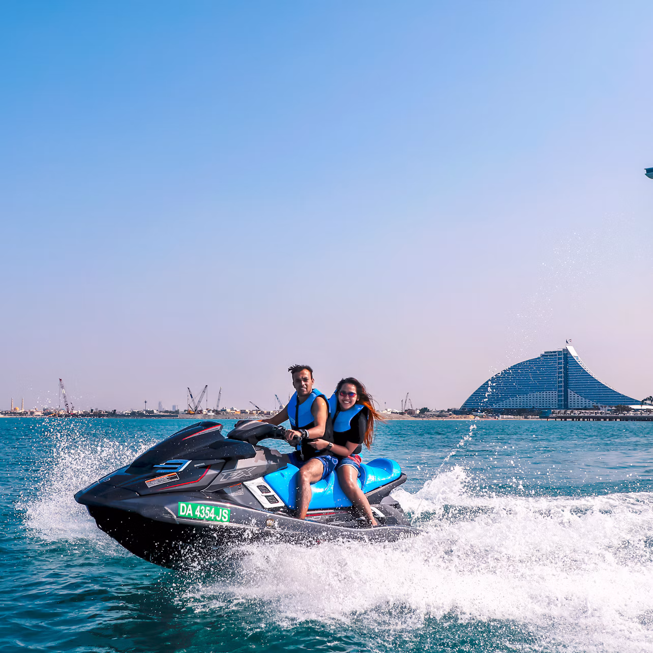 Two people riding a jet ski on open water, with a large, unique building and construction cranes visible in the background under a clear sky.