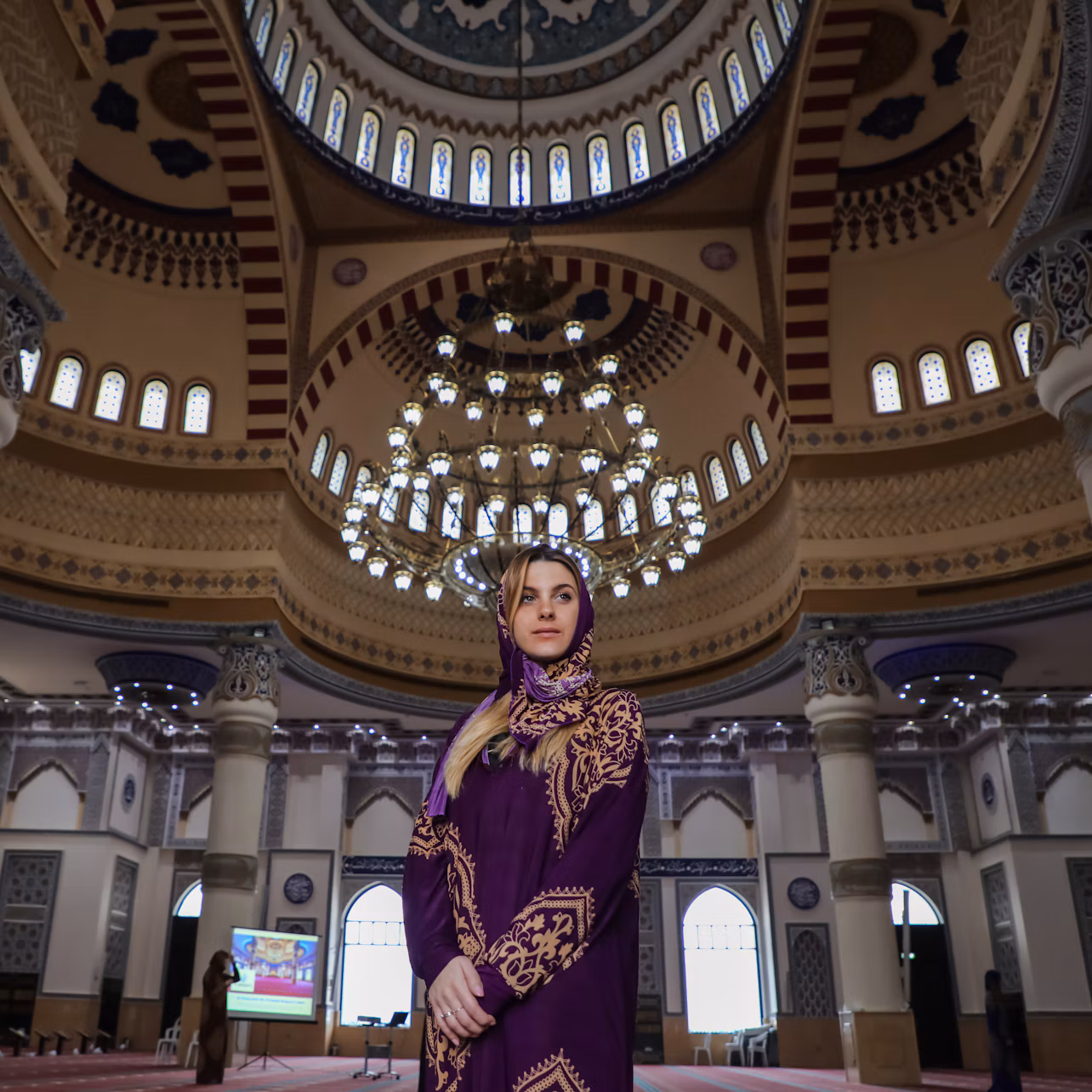 A woman in a purple headscarf stands inside an ornate mosque with a large chandelier and intricate architectural details.
