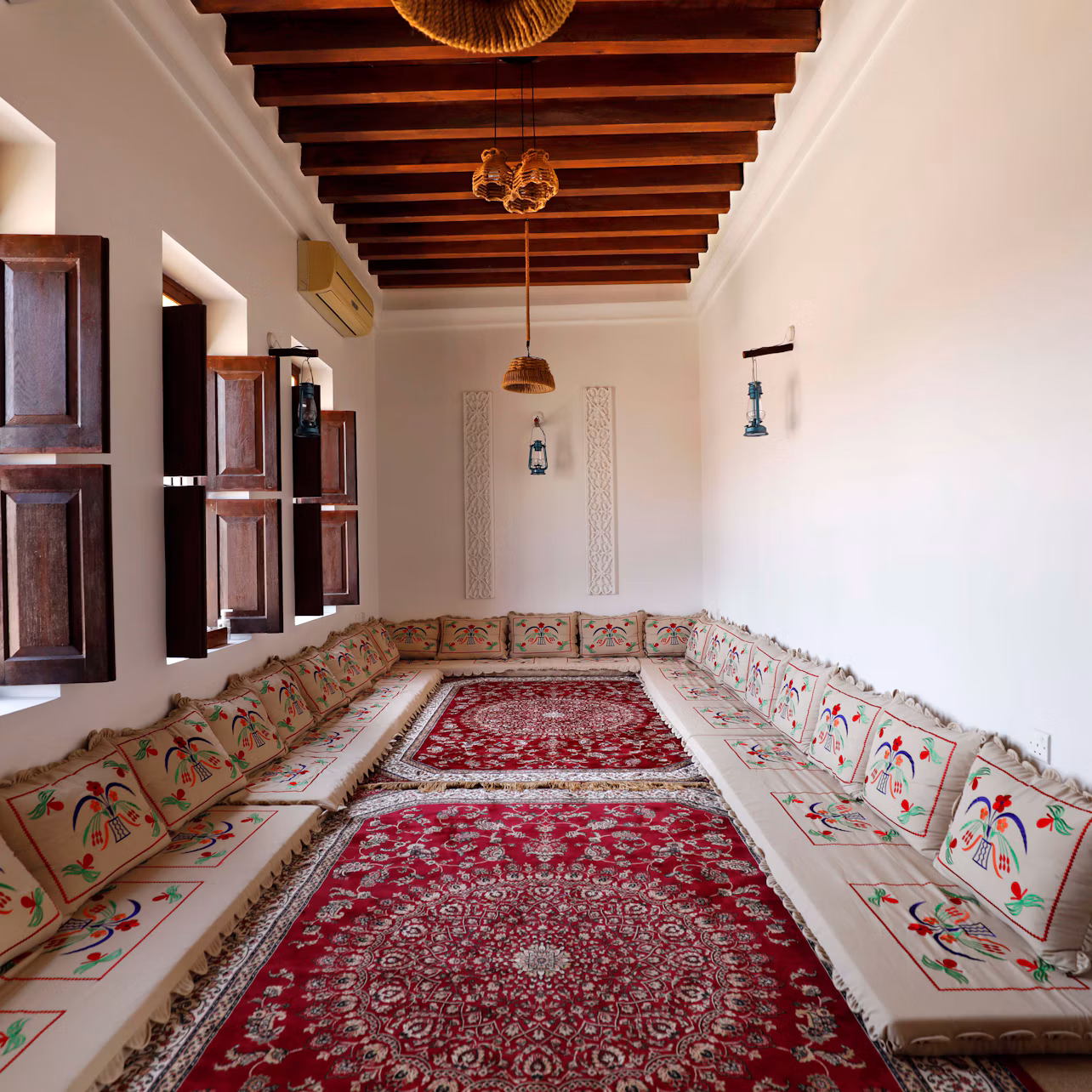 A long room with wooden ceiling beams, embroidered floor seating, red patterned rugs, and wooden shutters on the windows.