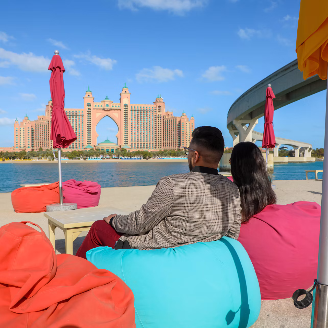 A man and woman sit on colorful bean bags near the water, with a large, ornate hotel in the background and some red umbrellas.