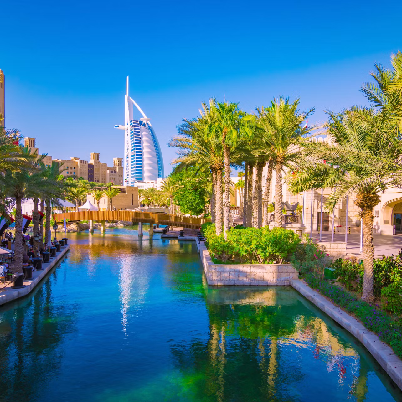 Modern skyscraper in the background, palm trees lining a blue canal with a bridge, and buildings in the foreground under a clear sky.