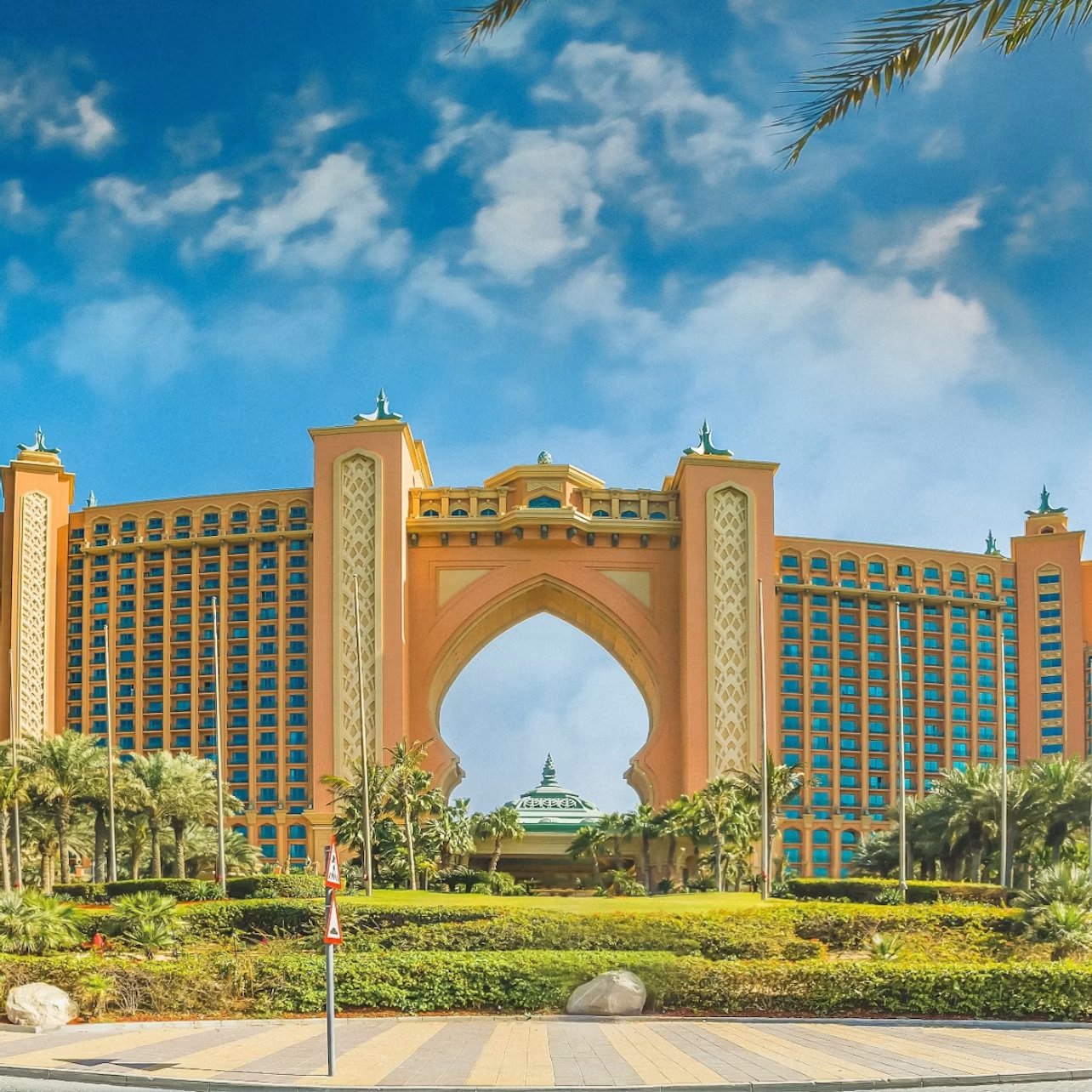 A large, luxury hotel with an archway in the center, surrounded by palm trees and lush landscaping, under a partly cloudy sky.