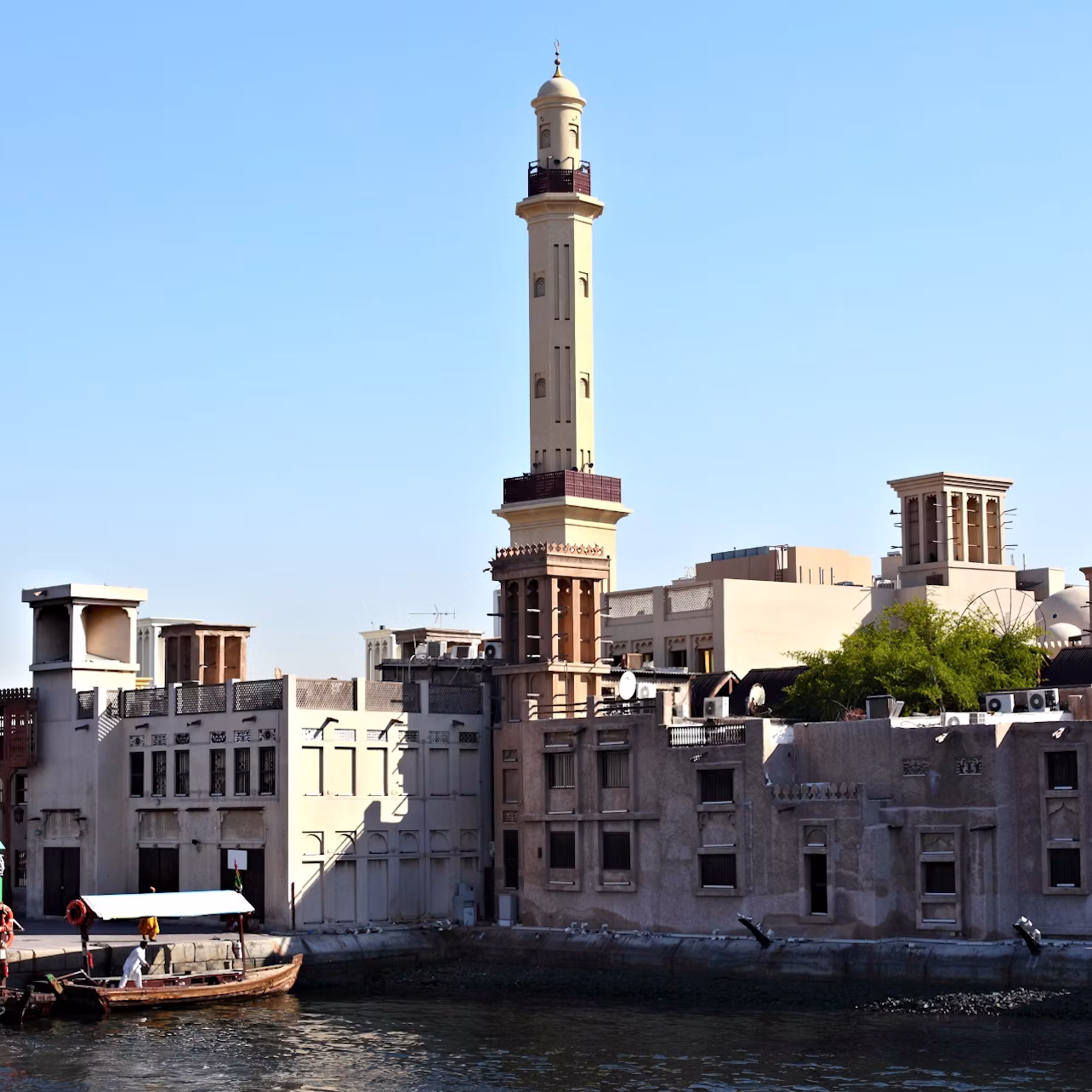 Traditional buildings and a tall minaret line a waterfront under a clear blue sky; a boat floats on the water in the foreground.