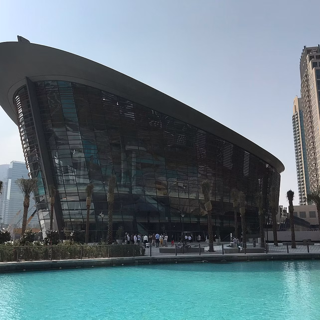 Modern glass building with a curved roof by a turquoise pool, surrounded by palm trees and skyscrapers in the background.