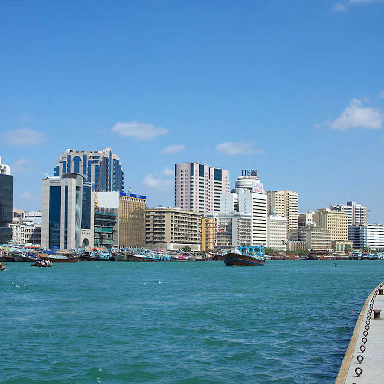 Skyline of a city with tall buildings along the waterfront, boats docked, and a few clouds in the blue sky.