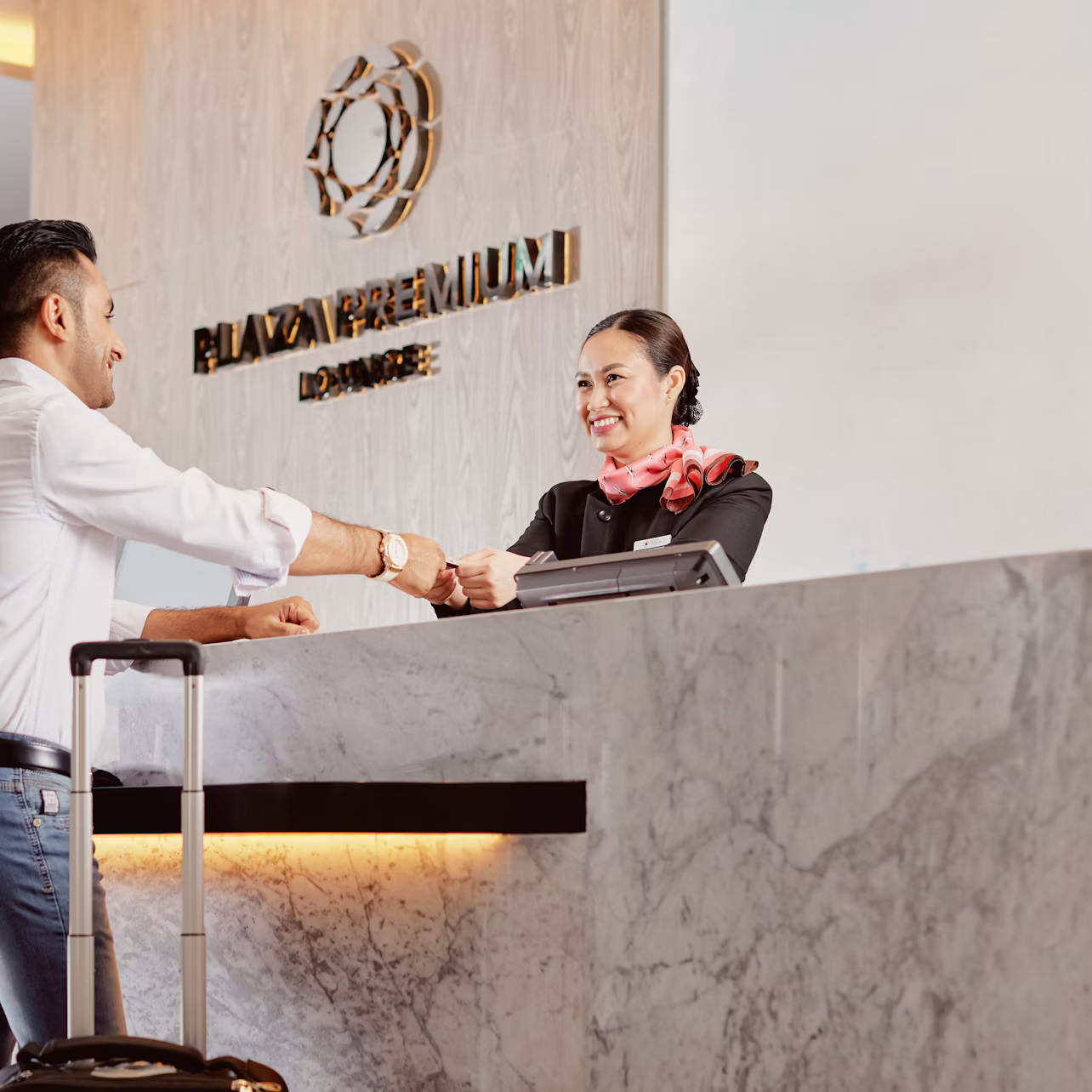 A man checks in at a reception desk while a smiling receptionist hands him a card. A suitcase stands beside him.