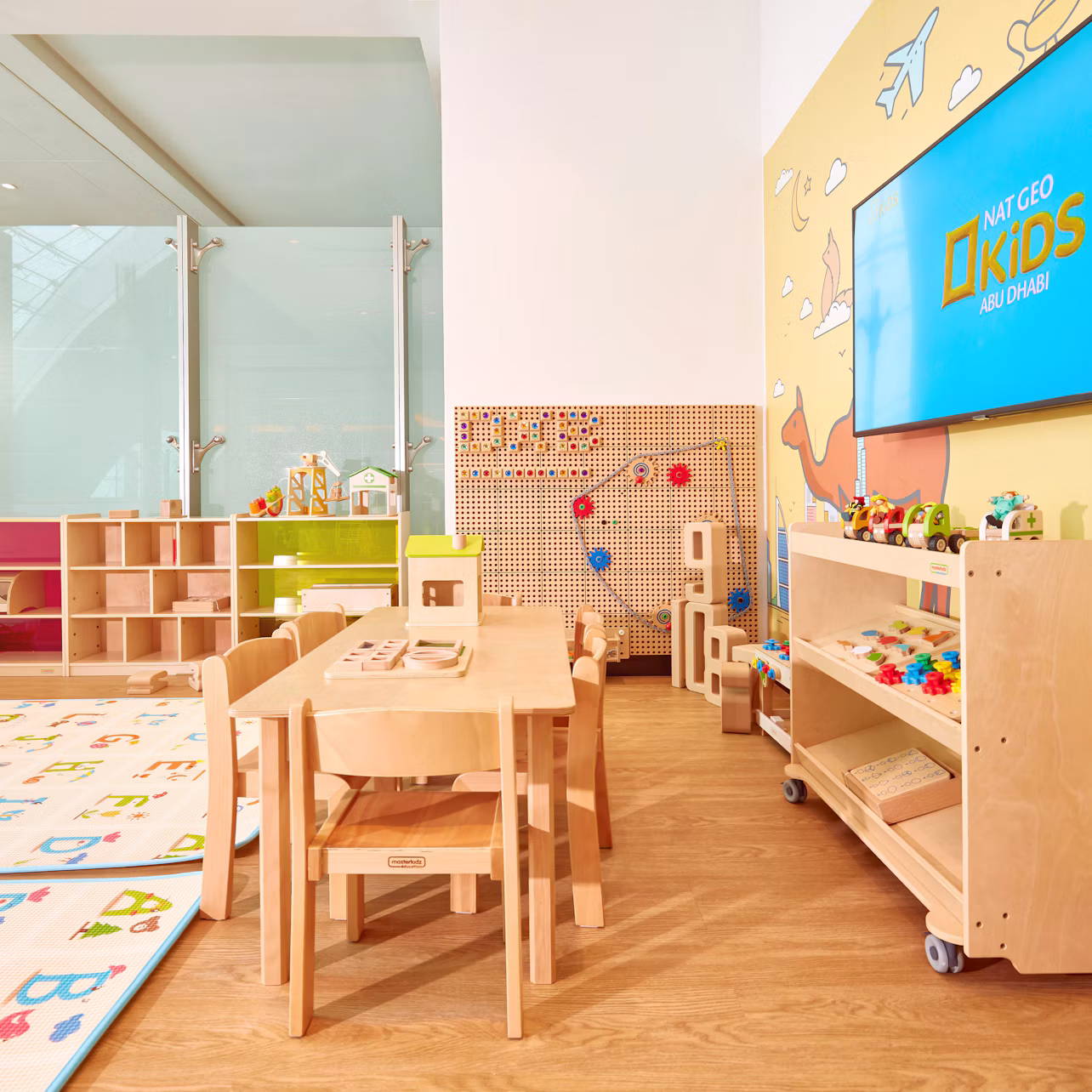 A colorful children's playroom with wooden tables, chairs, toys, shelves, and a TV displaying "Nat Geo Kids Abu Dhabi."