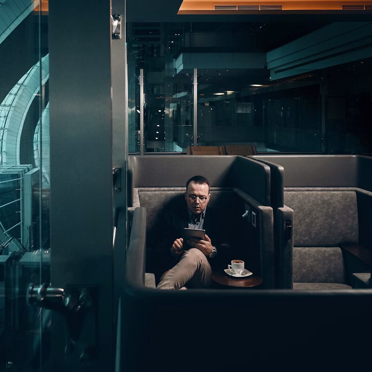A man reads a tablet while sitting in a private booth at a cafe, with a coffee cup and saucer on the table beside him.