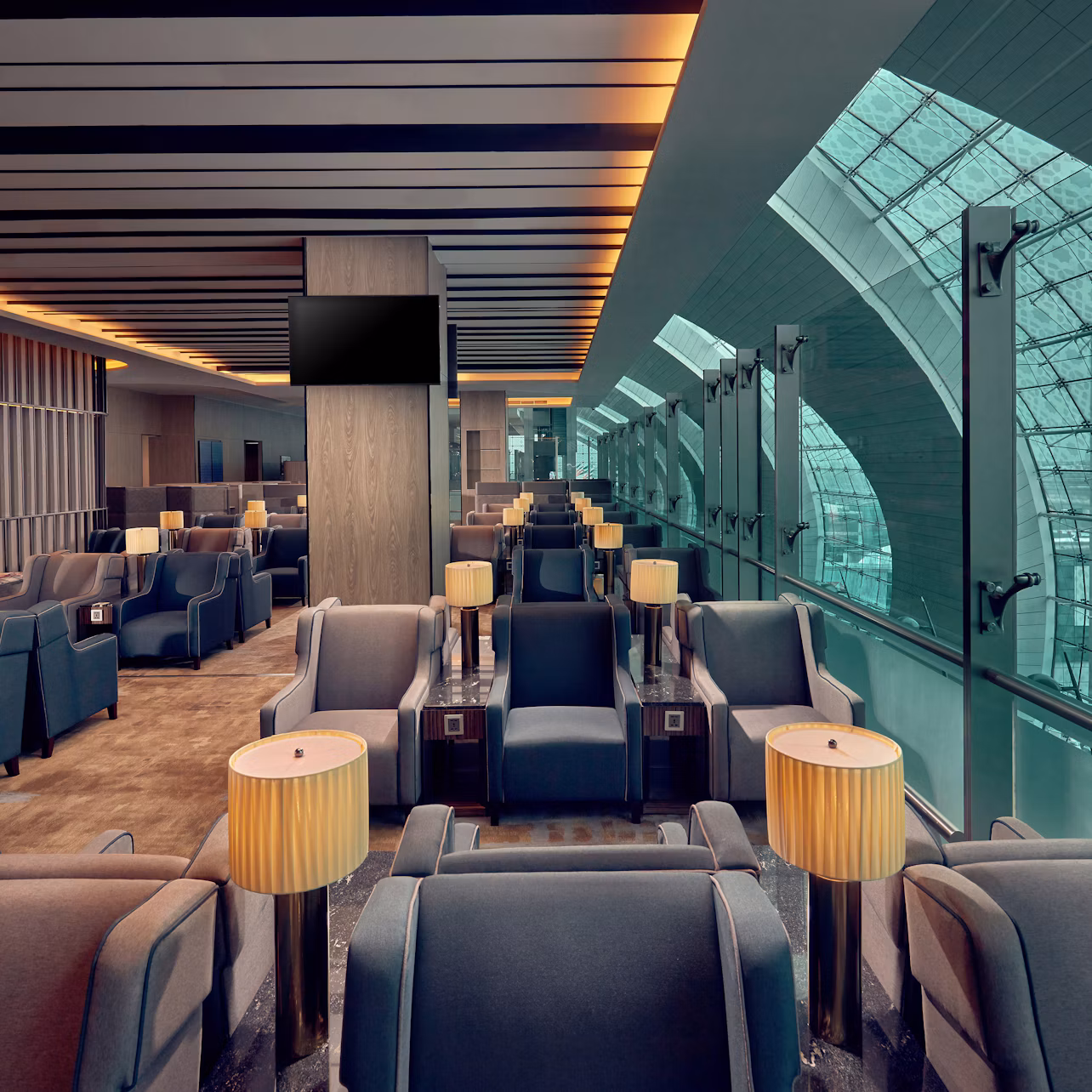 Modern airport lounge with grey seating, tables with lamps, and large windows showing an airport terminal.