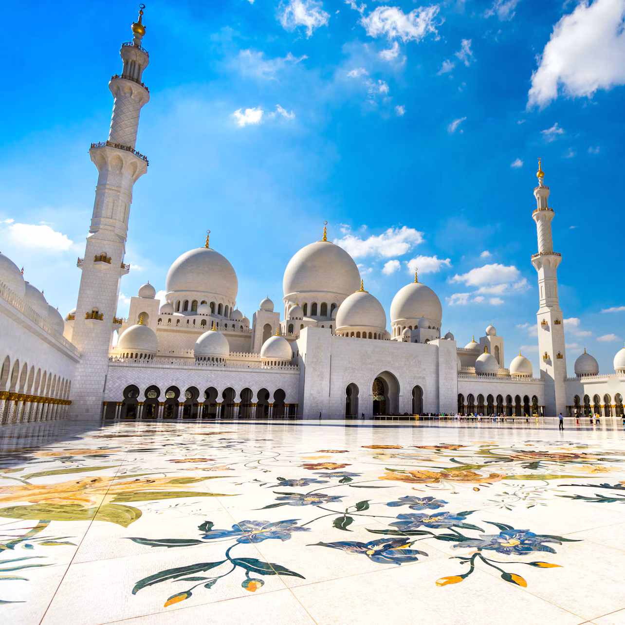 A white mosque with multiple domes, tall minarets, and an intricately tiled courtyard under a bright blue sky with clouds.