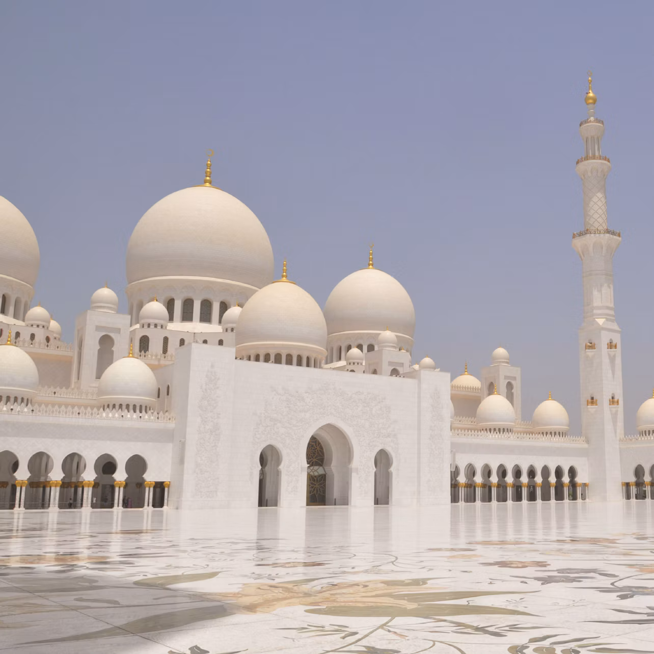 White mosque with large domes and minaret, intricate carvings, and spacious courtyard under clear blue sky.