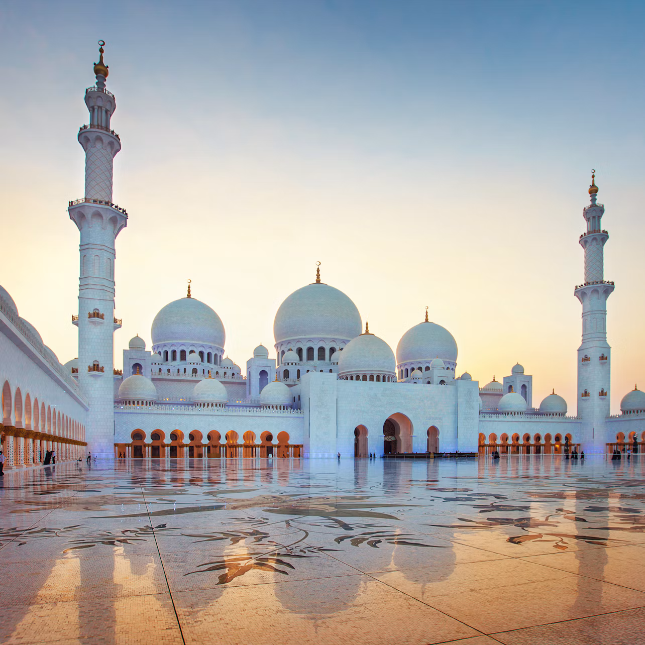 Grand mosque with multiple white domes and tall minarets at sunset, featuring intricate floral patterns on the courtyard floor.