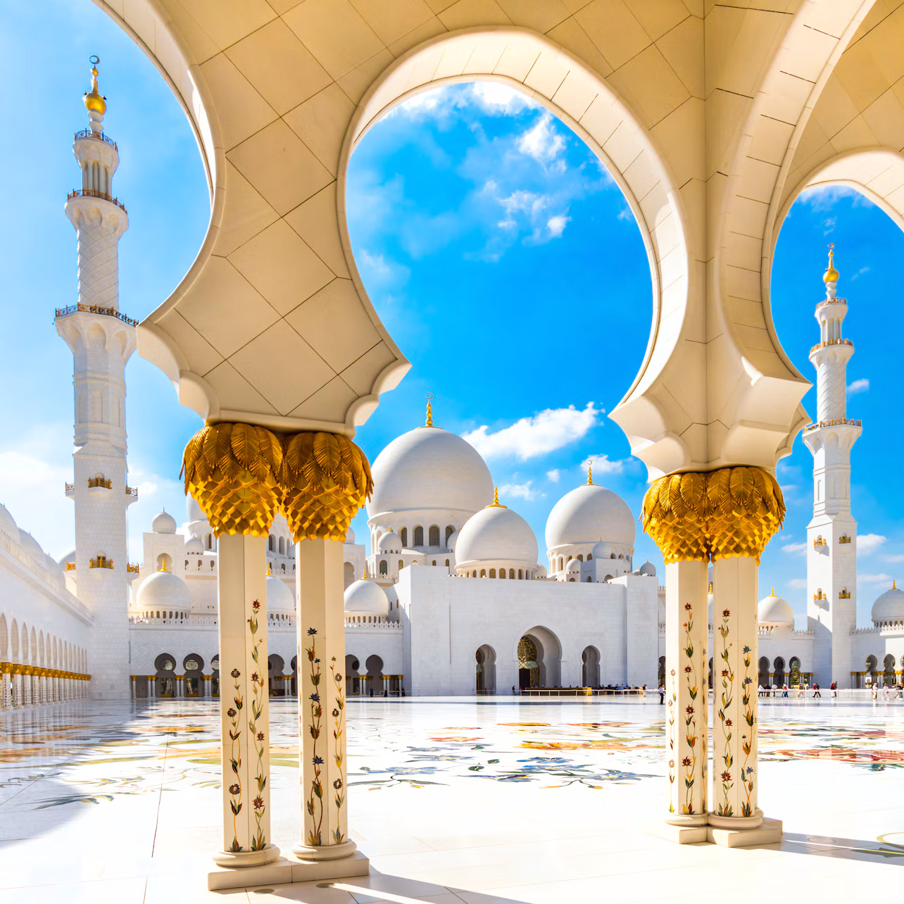 Intricate white and gold arches frame a courtyard with domes and minarets under a bright blue sky with some clouds.