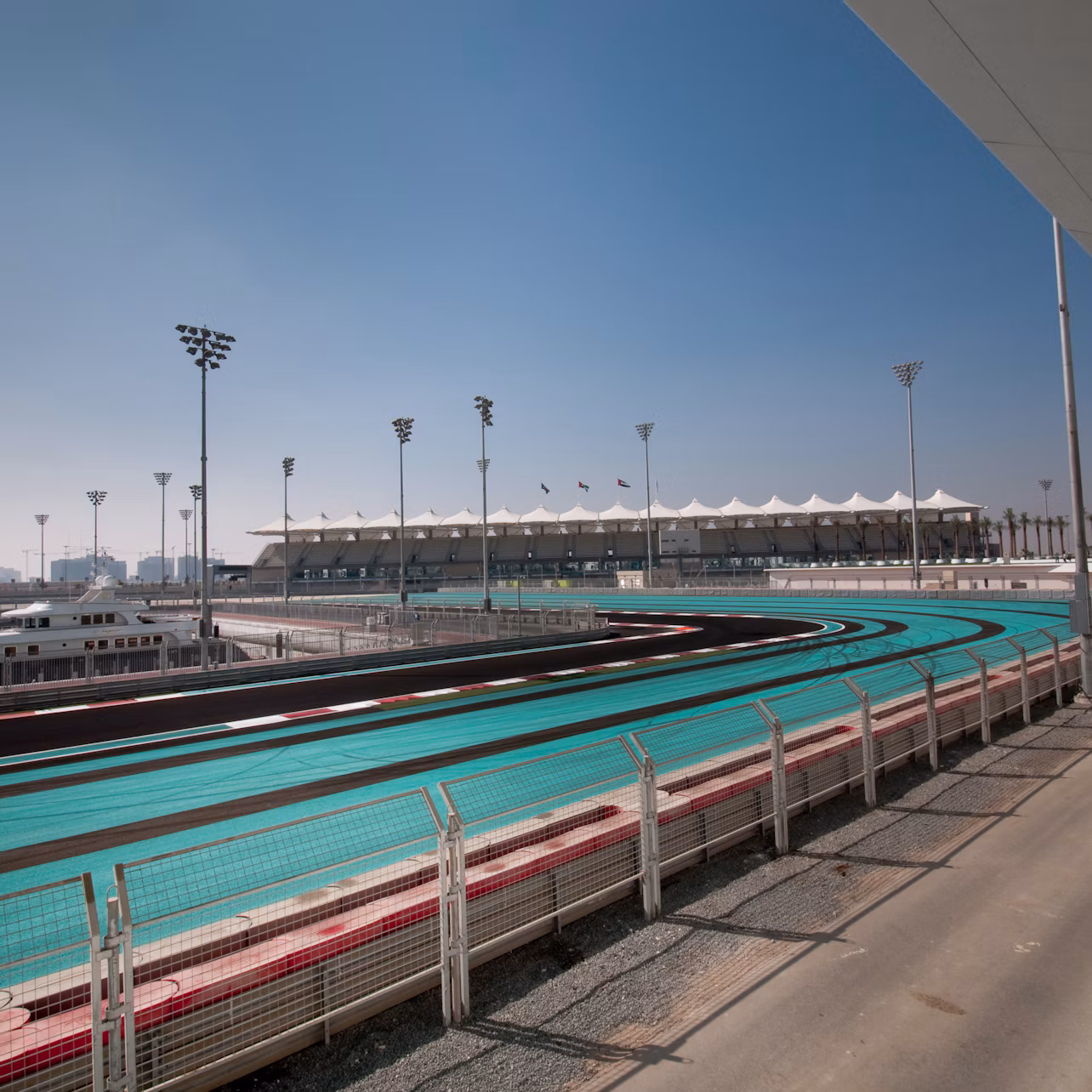 A curved racing track with turquoise and black lanes, under a clear blue sky, with a grandstand and floodlights in the background.