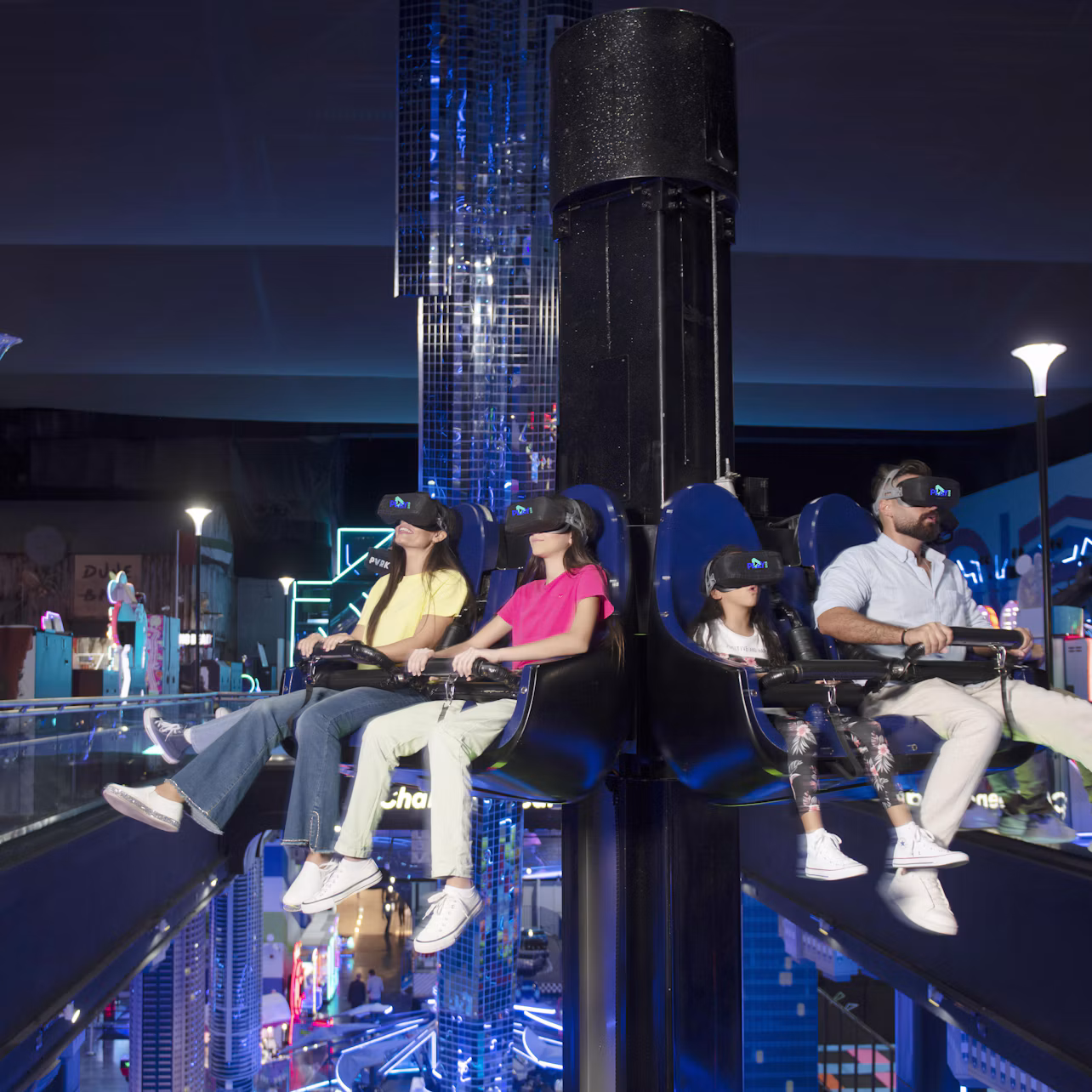 Four people wearing VR headsets are seated on a ride inside a futuristic, neon-lit indoor amusement park.