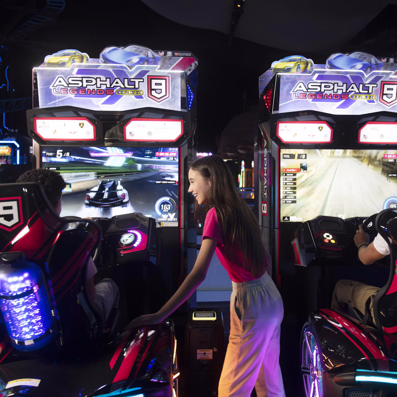 Two people play the racing game "Asphalt 9: Legends" at arcade machines while a third person watches.