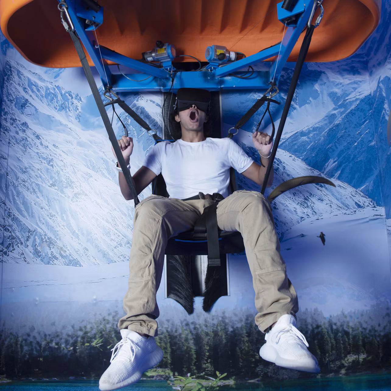 A man wearing a VR headset is seated on a suspended ride against a backdrop of snowy mountains, appearing excited.