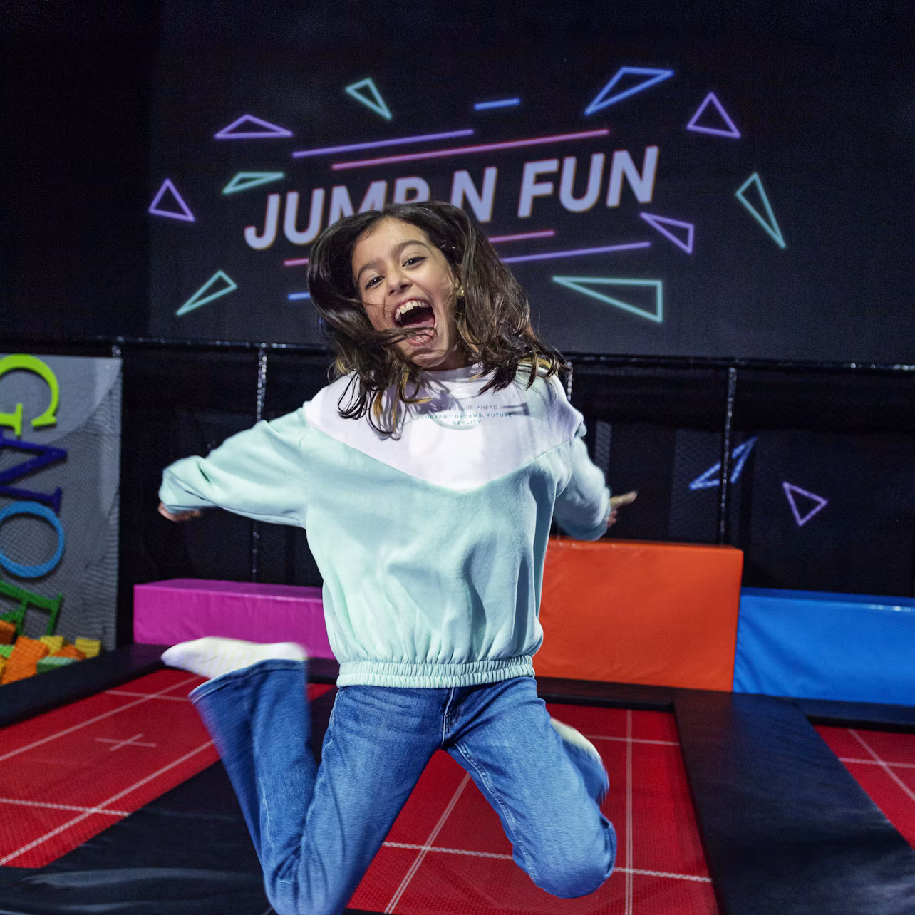 A child joyfully jumps on a trampoline at an indoor play area with colorful foam blocks and a sign that says "JUMP N FUN."