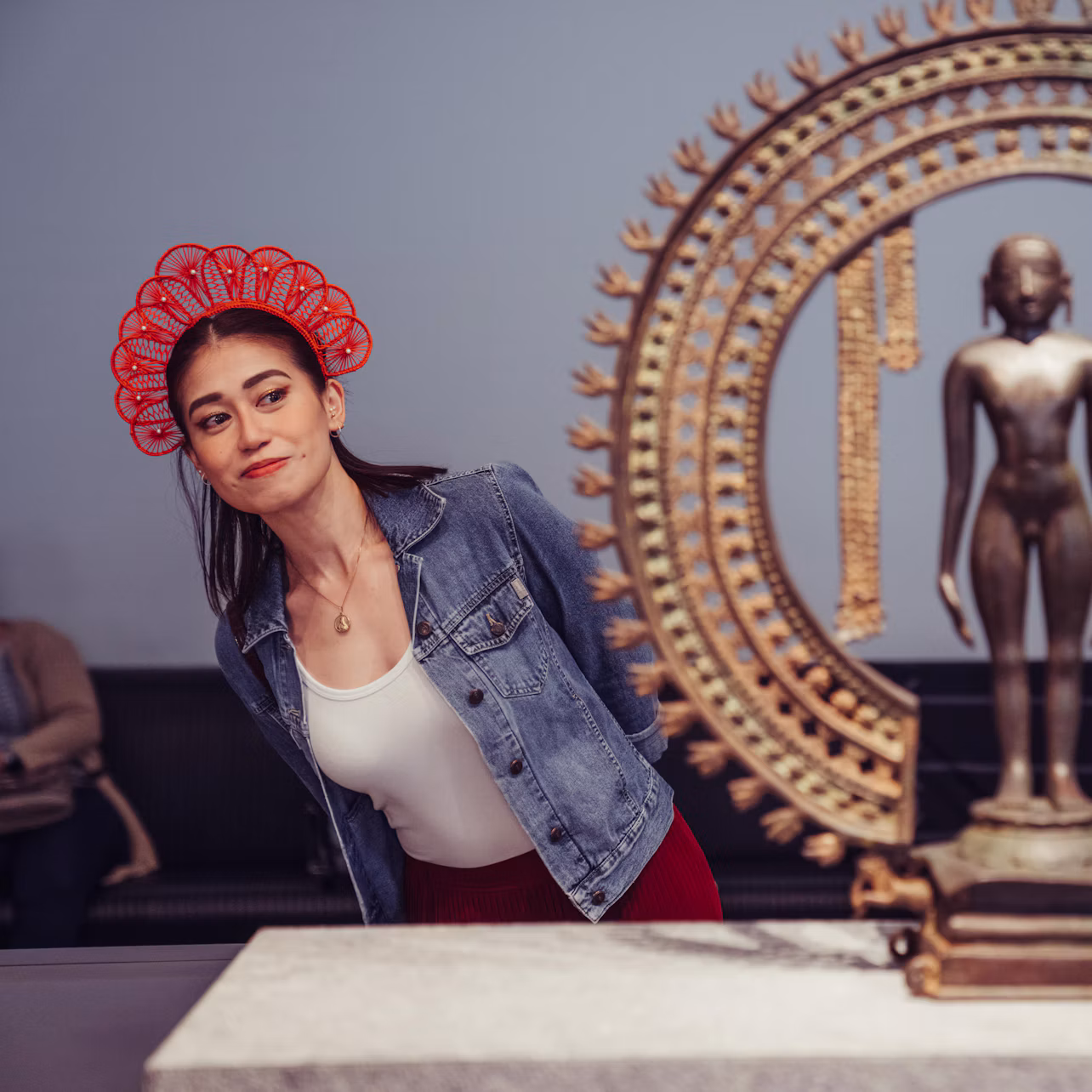 A woman in a red headdress and denim jacket leans to observe an ornate statue in a museum. A seated person is blurred in the background.