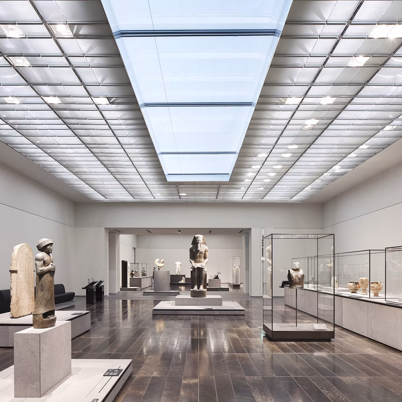A spacious museum gallery with ancient sculptures displayed on pedestals and in glass cases under a large skylight.