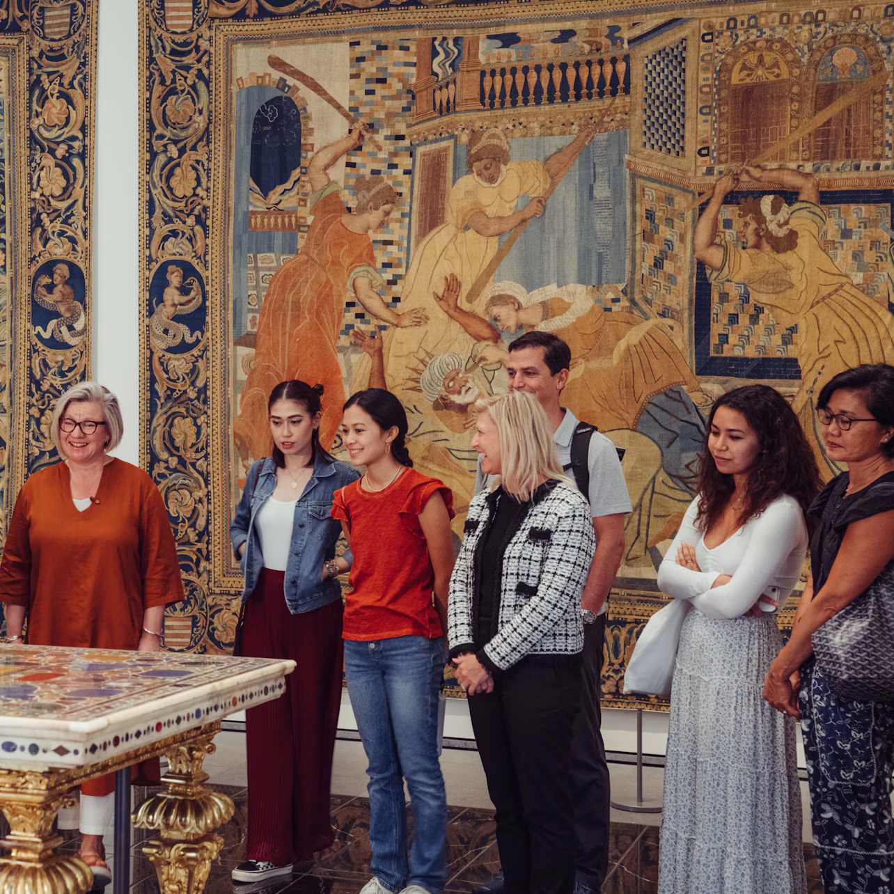 A group of people stand in an art gallery, observing a large, ornate tapestry with intricate designs and historical scenes.