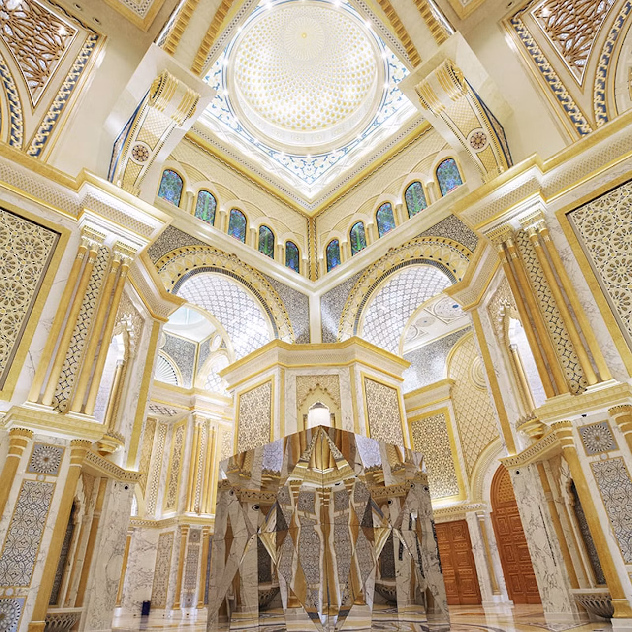 An opulent interior features ornate arches, columns, intricate patterns, a dome ceiling, and large mirrors reflecting the space.