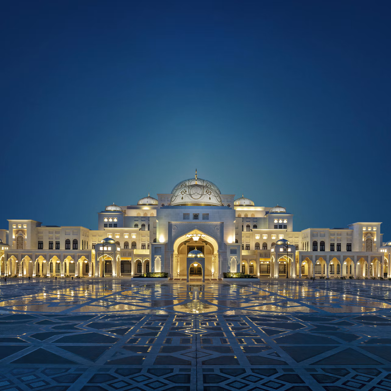 An illuminated grand palace with multiple arches and a central dome, reflected on the ornate pavement under a deep blue sky.