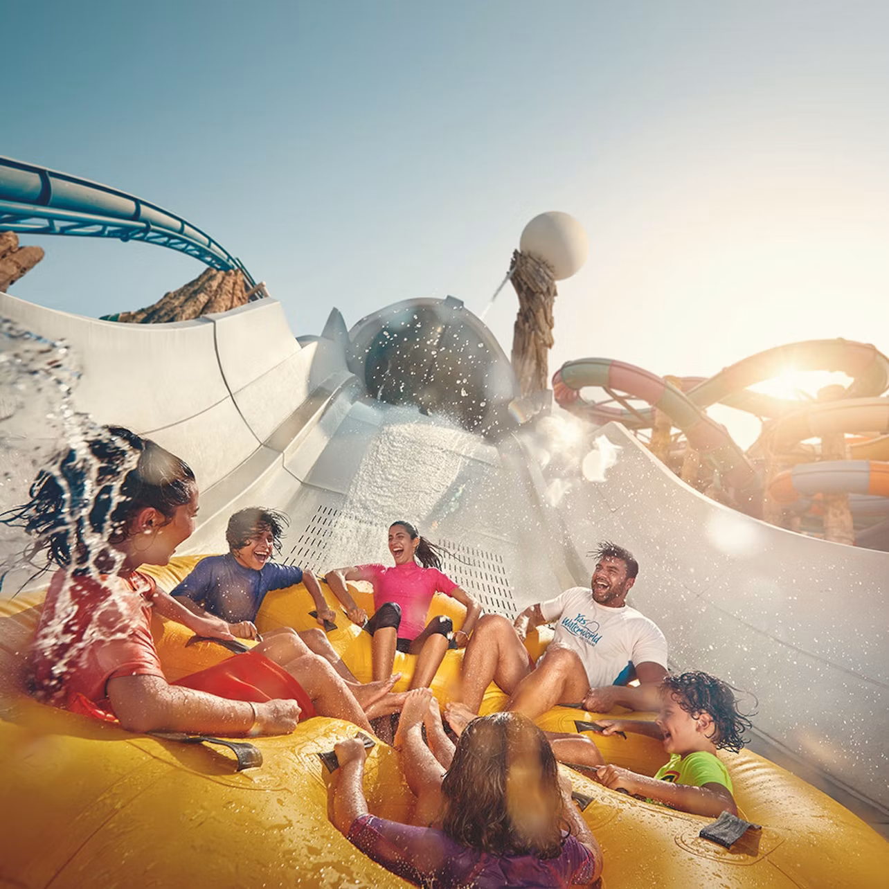People laughing and getting splashed while riding a large yellow raft on a water slide under a sunny sky.