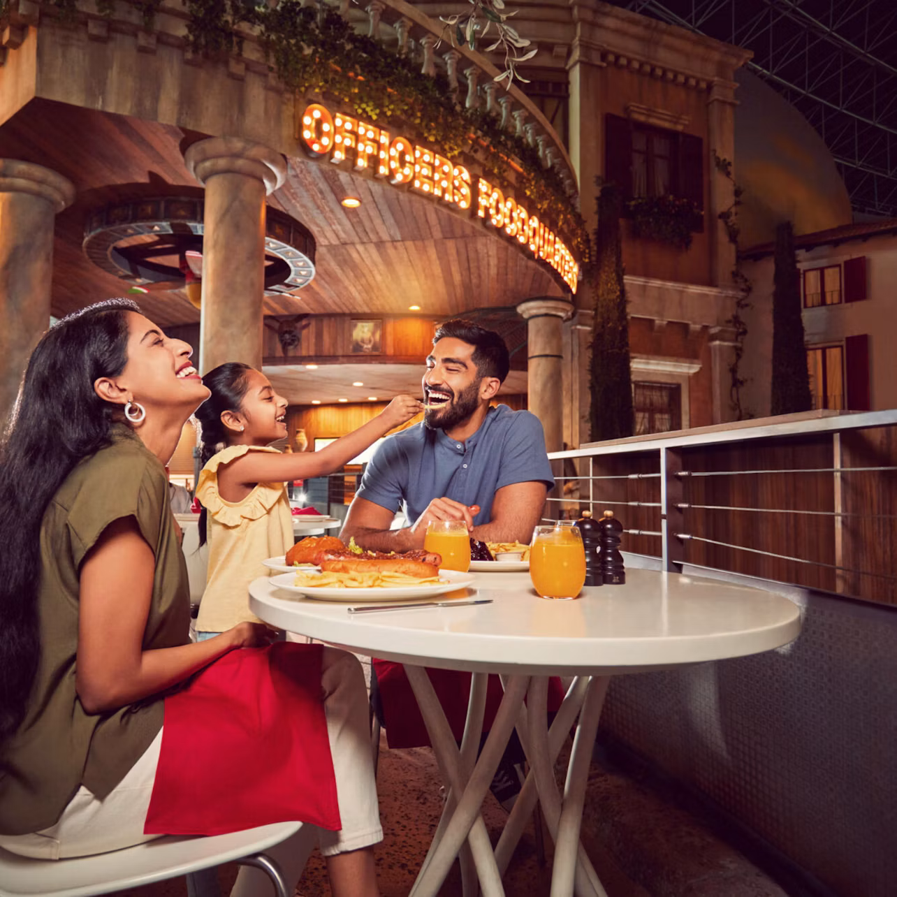 A family of three enjoys a meal together at an outdoor restaurant, smiling and sharing food on a well-lit terrace.
