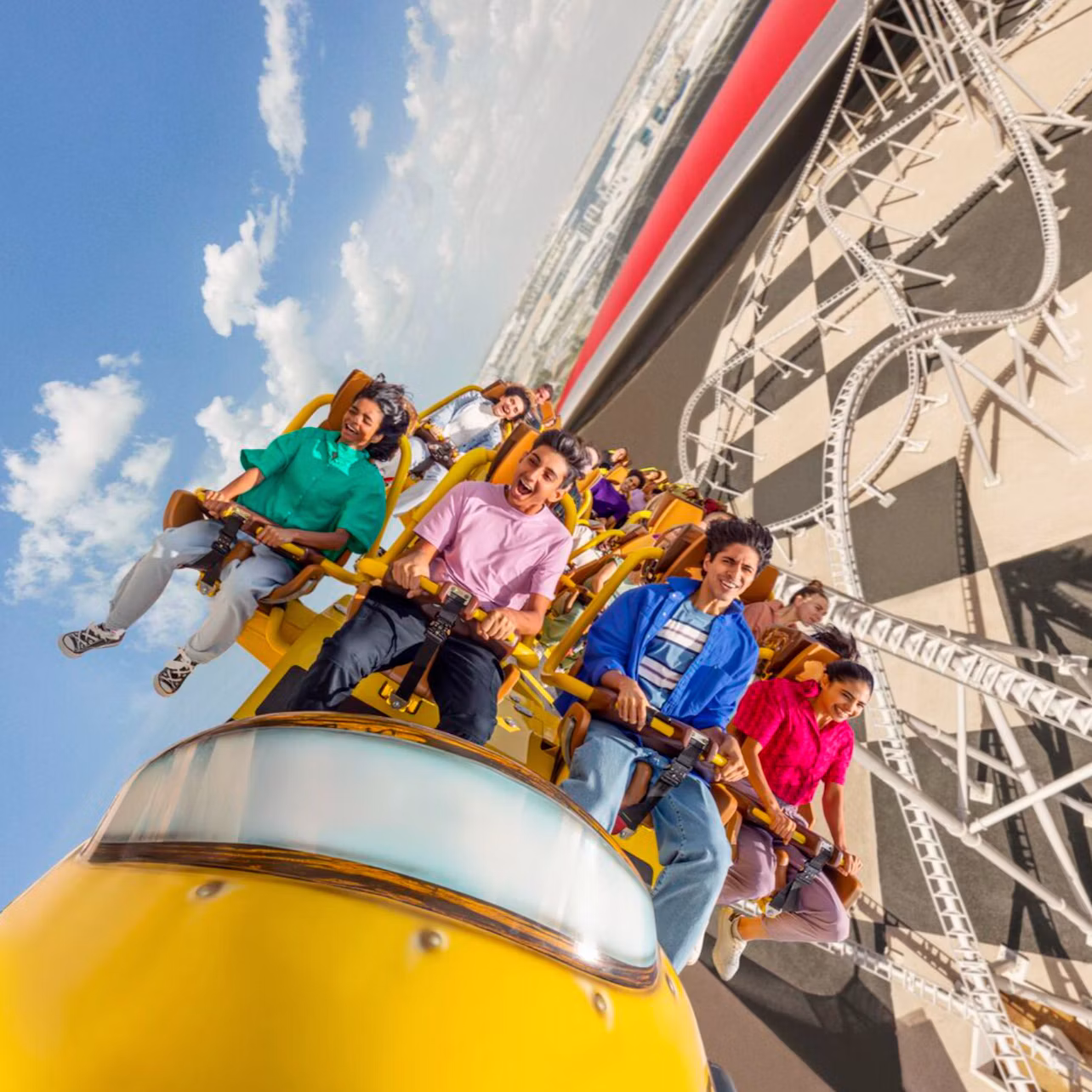 People riding a yellow roller coaster with visible tracks and a building in the background under a blue sky.