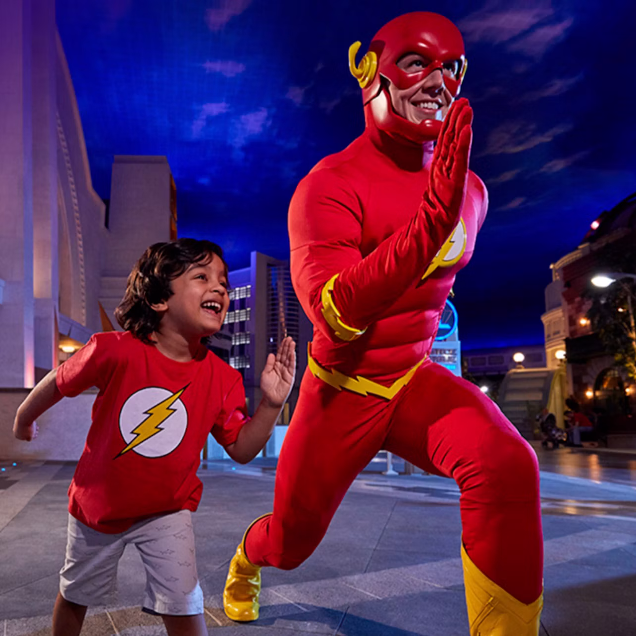 A child wearing a Flash-themed shirt poses joyfully next to a person in a Flash superhero costume in an urban evening setting.