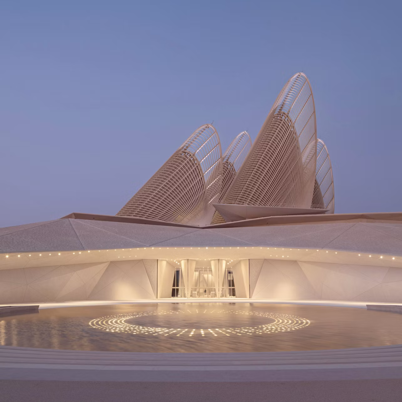 Modern architectural structure with curved, sail-like elements under a twilight sky, featuring a circular lit pattern in front.