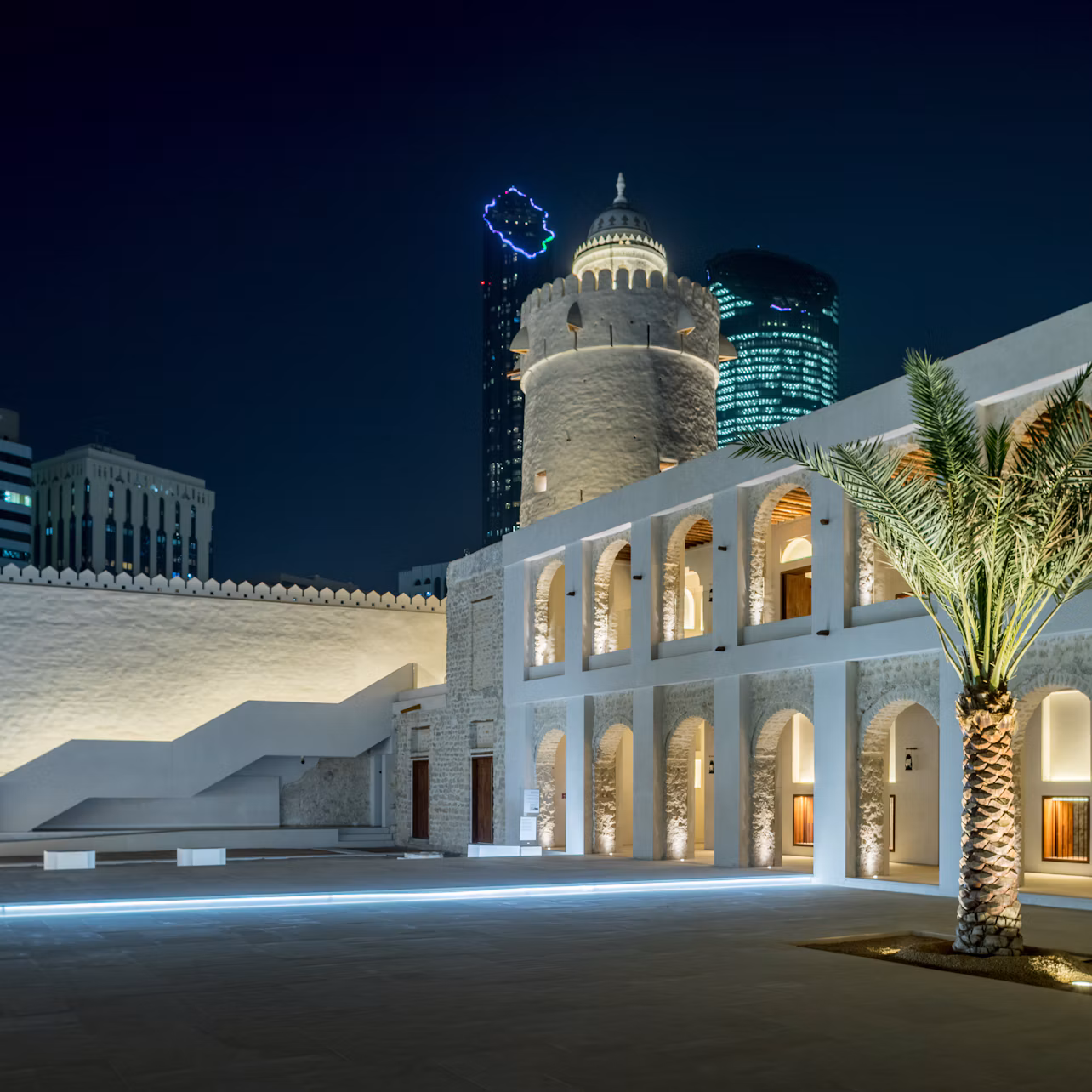 Stone building with arched doorways and a central dome lit at night, palm tree in the foreground, modern buildings in the background.