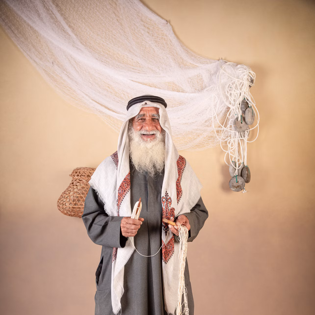 An elderly man with a long white beard, wearing traditional attire, holds fishing net repair tools against a beige background with a draped net.