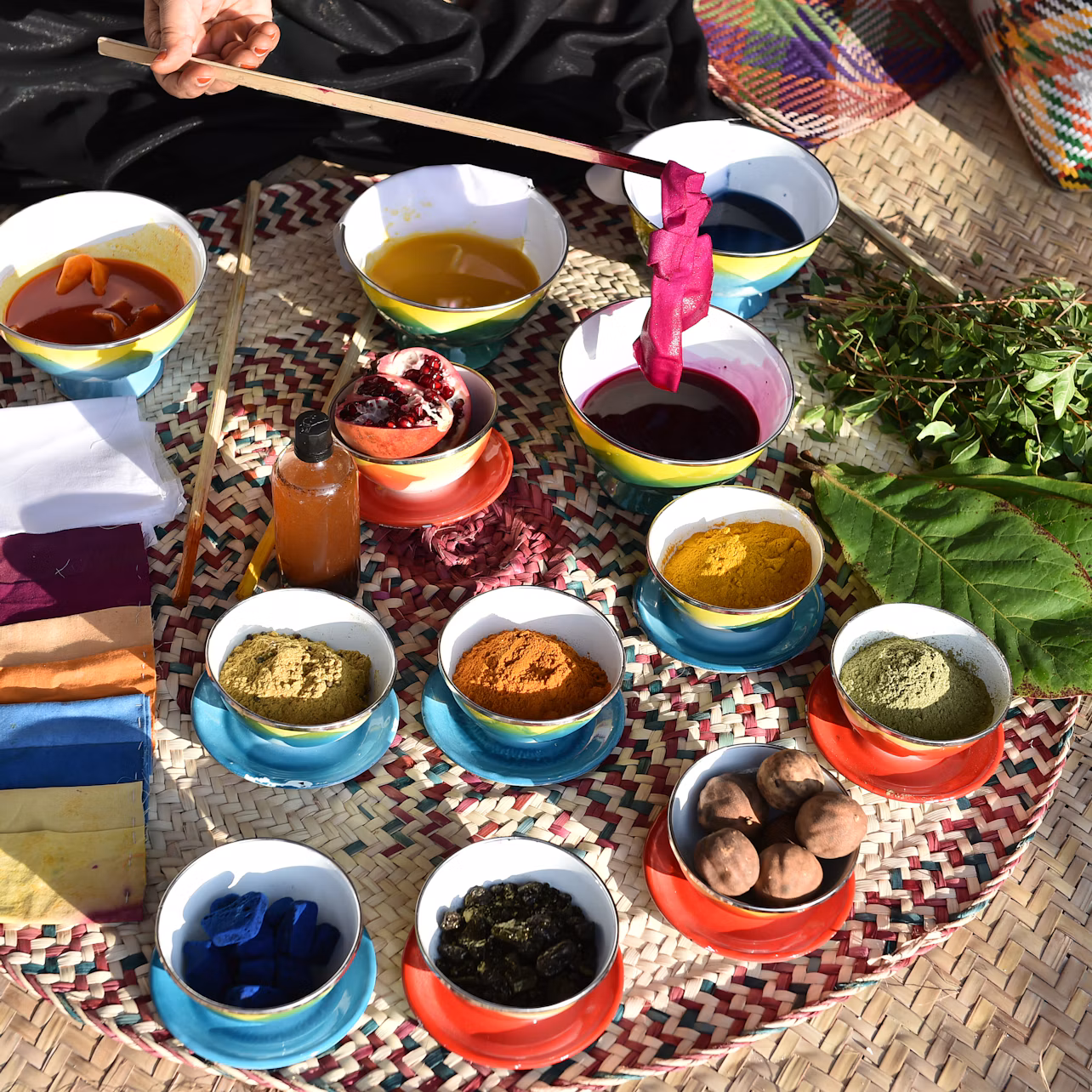 A collection of colorful natural dye ingredients and pigments in bowls, a person's hand holding a stick with dyed fabric.