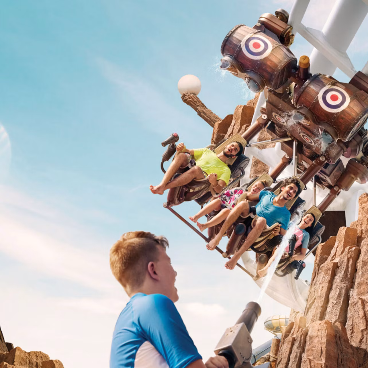 People riding a swinging theme park ride shaped like barrels, with a child aiming a water cannon at them.