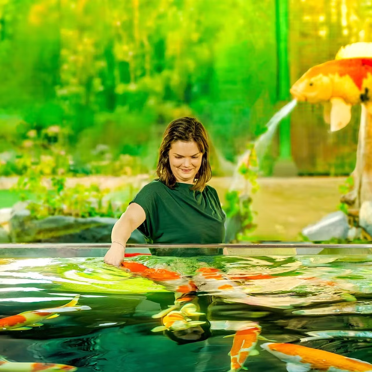 A woman interacts with koi fish in a pond surrounded by greenery and a decorative sculpture of a fish.